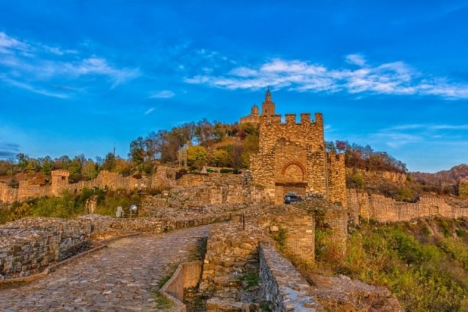 A stone pathway leads up to the Tsarevets fortress in Veliko Tarnovo, Bulgaria, with its ancient walls and towers against a bright blue sky.